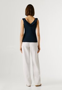 Marinblå topp med volangkantad V-ringning, figursydd överdel, bärs med vita vida byxor. Tyget verkar slätt, minimalistisk design.