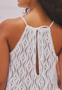 Haut sans manches blanc avec un motif texturé et perforé, doté d'un lien dans le dos et d'une découpe en forme de trou de serrure pour l'aération.