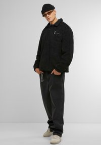 Pullover in pile nero con colletto, bottoni frontali e logo ricamato. Abbinato a jeans neri a vestibilità ampia e sneakers bianche.