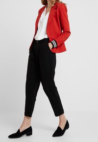 Punainen Blazer, jossa on musta-valkoiset raidalliset hihansuut, valkoinen paita, mustat pleatatut housut ja mustat mokkanahkakengät. Moderni istuvuus ja puhdas linja.