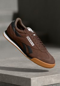 Sneakers din suede maro cu accente negre, etichetă albă cu logo și talpă din cauciuc de tip gumă. Dispune de închidere cu șireturi și design cu vârful rotund.