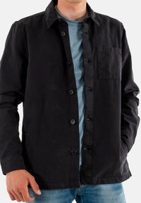 Zwarte getailleerde jas met een gestructureerde kraag, franjes en een zachte textuur, gedragen over een blauwe ronde hals shirt.