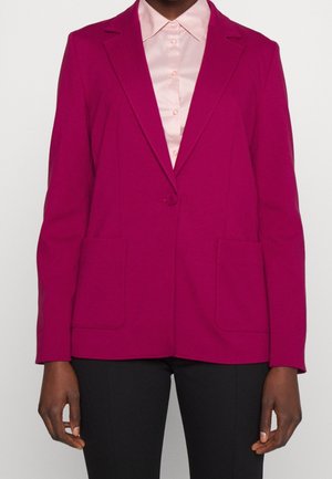 Person iført en lys magenta blazer med to forlommer over en lys lyserød skjorte med knapper og sorte bukser.