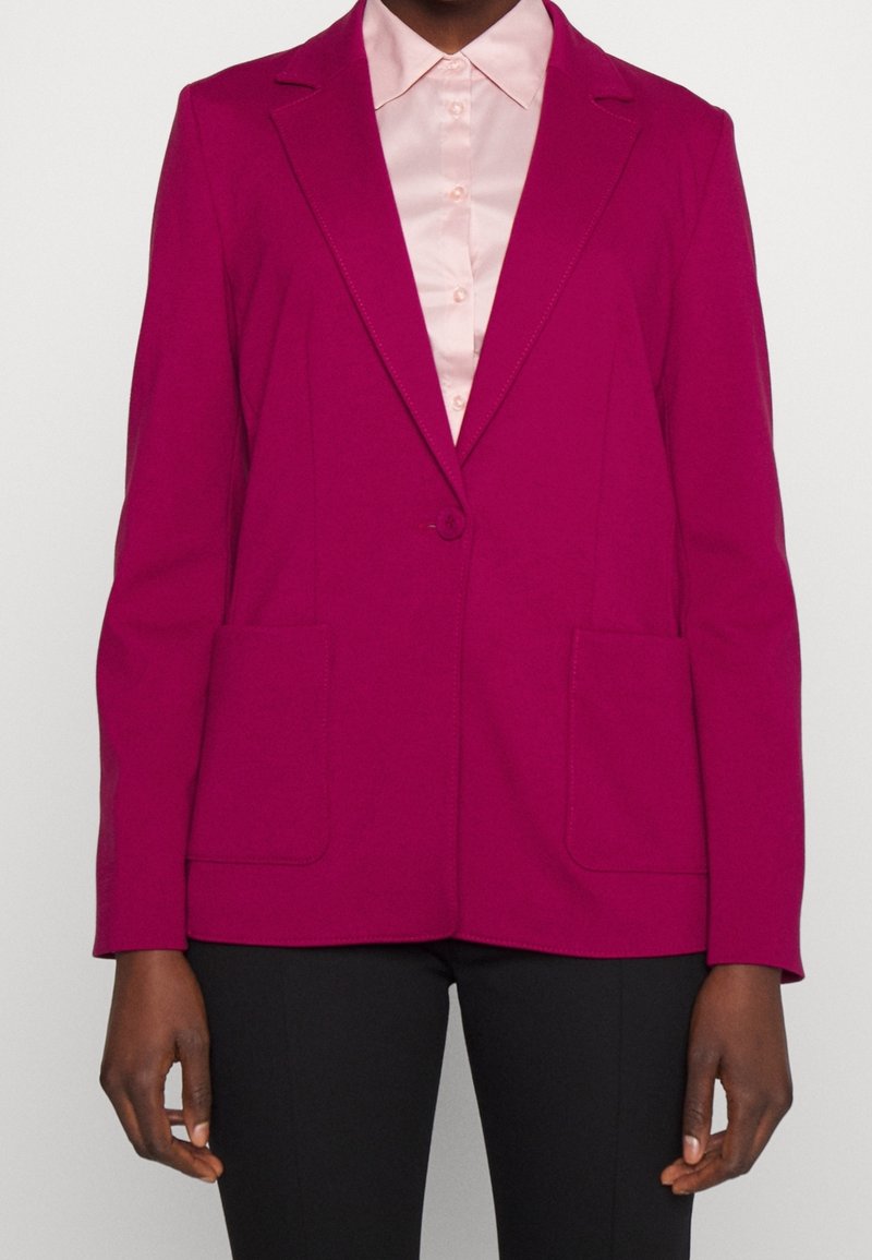 Personne portant un blazer magenta vif avec deux poches avant sur une chemise rose pâle boutonnée et un pantalon noir.