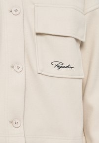 Light beige fleece shirt jacket features a button closure, one large pocket, and embroidered "Pegador" logo in black. Soft texture.