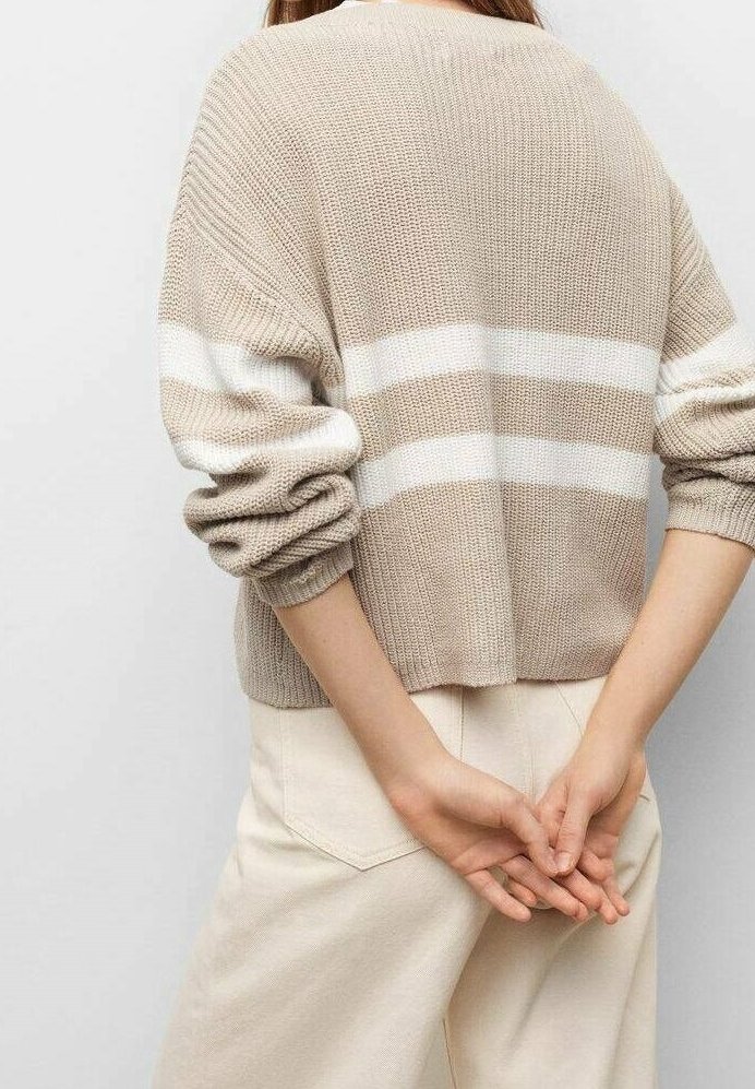 Personne portant un pull en tricot beige avec des rayures horizontales blanches et un pantalon clair, vue de dos avec les mains jointes derrière.
