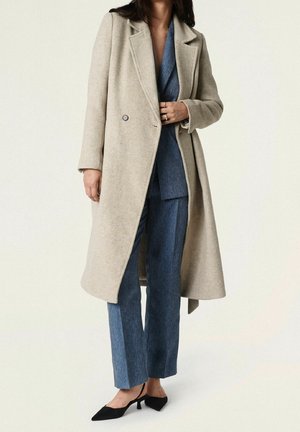 Woman wearing a beige long coat over blue tailored pants and blazer, paired with black pointed slingback heels against a plain background.