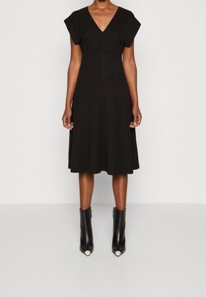 Mujer con vestido negro hasta la rodilla, de manga corta y escote en V, combinado con botines negros con puntas de color claro.