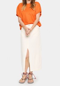 Oranssi lyhythihainen paita, joka on yhdistetty kermaiseen, polvimittaisiin hameeseen, jossa on etureikä. Asusteena moniväriset, remmillä varustetut sandaalit.