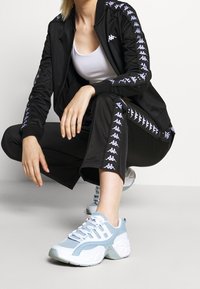 Svart träningsjacka med vita logodetaljer, matchad med svarta byxor med logotyper. Ljust blå sneakers med vita detaljer.