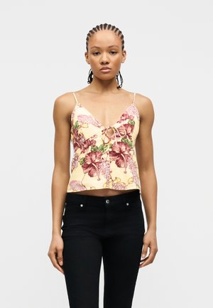 Νέα γυναίκα με πλεξούδες φορώντας κίτρινη φλοράλ καμισόλη και μαύρο παντελόνι, στέκεται μπροστά σε ένα λευκό φόντο.