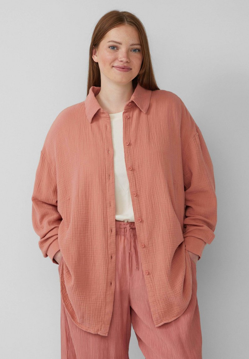 Chemise à boutons corail à coupe ample avec un tissu texturé, associée à un pantalon plissé assorti. Présente un col et des poches latérales.