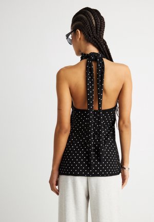 Γυναίκα με πλεγμένα μαλλιά που φοράει μαύρο τοπ με λευκές βούλες και δέσιμο στον λαιμό, σε συνδυασμό με ανοιχτόχρωμο παντελόνι, μπροστά σε λευκό φόντο.