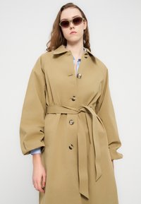Καμηλό παλτό τραντζ με ζώνη στη μέση, μεγάλα κουμπιά και φαρδιά μανίκια. Ορατό γαλάζιο πουκάμισο από κάτω, σε απλό φόντο.
