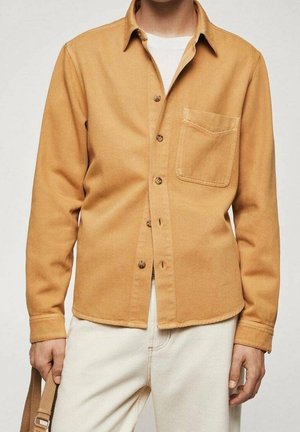 Homme portant une chemise boutonnée jaune moutarde par-dessus un t-shirt blanc et un pantalon couleur crème, tenant une lanière beige dans la main gauche.