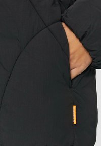Zwarte pufferjack met gestikte ruitpatroon en een zijzak. Heeft een gestructureerde stof en een kleine oranje tag aan de zoom.
