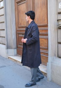 Mand i mørk lang frakke justerer ærmerne, mens han står på en fortov nær en stor trædør i en urban setting.