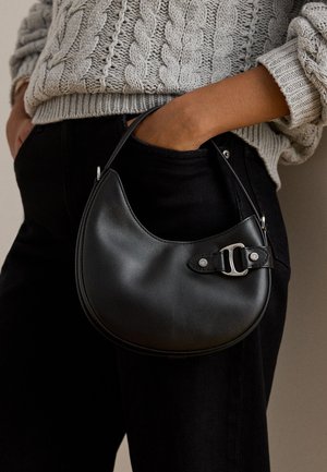 Άτομο με γκρι πουλόβερ πλεκτό καλώδιο και μαύρο παντελόνι κρατά μικρή μαύρη δερμάτινη τσάντα μισοφέγγαρο με λεπτομέρεια ασημένιας αγκράφας.