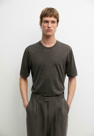 Giovane uomo con capelli castani corti che indossa una camicia marrone scuro a maniche corte e pantaloni abbinati, in piedi con le mani in tasca davanti a un muro bianco.