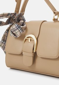 Beige leren handtas met een gestructureerd ontwerp, gouden hardware en een decoratieve geprinte sjaal geknoopt aan de handgreep.