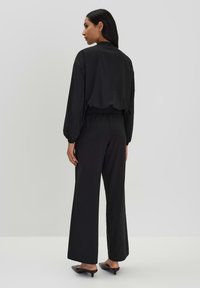Schwarzer, locker sitzender Jumpsuit mit langen Ärmeln und elastischem Bund; kombiniert mit eleganten schwarzen Blockabsatzschuhen. Glatte Stoffstruktur.