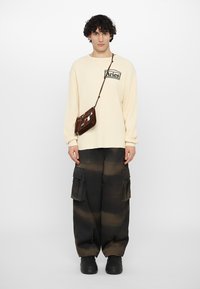 T-shirt beige à manches longues côtelé avec un graphique noir, associé à un pantalon cargo noir avec poches et un sac bandoulière marron. Chaussures noires.