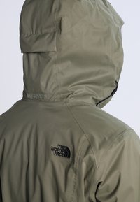 Olivgrüne wasserdichte Jacke mit Kapuze, versehen mit einem schwarzen Logo auf der Rückseite, strukturierten Oberflächen und verstellbarem Kordelzug in der Nähe des Gesichts.