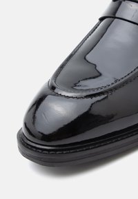 Fekete lakkozott bőr mokaszinok lekerekített orral, varrott részletekkel és sima felülettel. Gumi talp, enyhe mintázattal.