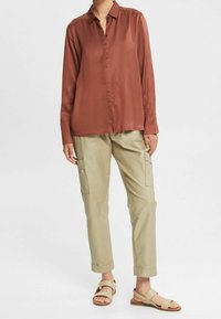 Vrouw in een roestkleurige blouse met lange mouwen, beige cargobroek met omgeslagen zoom en beige sandalen met open tenen, staand op een witte achtergrond.