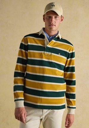 Man wearing cream cap and green, yellow, and white striped long-sleeve rugby shirt with white collar and beige pants.