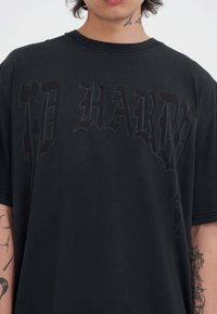 Czarna, oversize T-shirt z ciemnym graficznym tekstem, okrągłym dekoltem i krótkimi rękawami. Materiał wydaje się miękki o gładkiej fakturze.