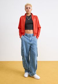 Giacca da track rossa con zip, crop top nero, pantaloni cargo blu con tasche laterali, sneakers bianche; la modella indossa occhiali da sole rosa.