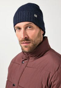 Dark blue ribbed knit beanie paired with a maroon padded jacket featuring black snap buttons; close-up of face with light beard.