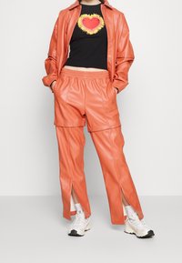 Completo in finta pelle arancione con giacca e pantaloni. La giacca ha una vestibilità comoda, mentre i pantaloni presentano dettagli con bottoni a pressione e spacchi laterali. Maglietta nera con grafica sotto.