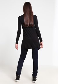 Tunica nera a maniche lunghe con orlo curvo, abbinata a jeans skinny scuri e sneakers alte nere. Tessuto morbido, design minimalista.