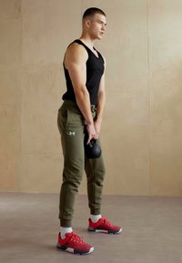 Schwarzes ärmelloses Shirt, olivgrüne Jogginghose, rote Sportschuhe mit weißen Sohlen; hält eine schwarze Kettlebell und steht auf einem hellen Boden.