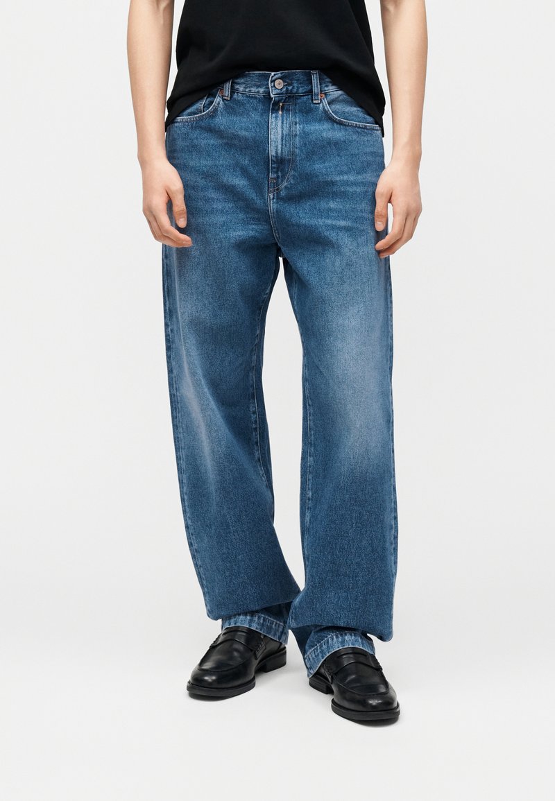 Άτομο που φοράει άνετα μπλε τζιν παντελόνια σε συνδυασμό με μαύρα δερμάτινα μοκασίνια και μαύρο πουκάμισο μπουκαρισμένο μέσα, δείχνεται από τη μέση μέχρι τα πόδια.