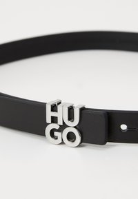 Cintura in pelle nera con lettere in metallo argentato che formano la parola "HUGO". Presenta una texture liscia, lunghezza regolabile e un design minimalista.