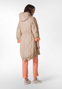 Cazadora larga acolchada beige con capucha, cintura ceñida y mangas con puños color coral, combinada con pantalones coral y zapatillas multicolores.