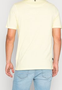 Světlounce žluté tričko s krátkými rukávy vyrobené z měkké bavlny, s kulatým výstřihem a malým černým logem na zádech. Nosit s modrými džínami.