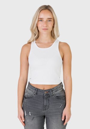 Donna bionda con capelli fino alle spalle che indossa un top corto bianco senza maniche e jeans grigi slavati strappati, in piedi davanti a uno sfondo semplice.