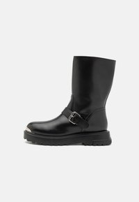 Melna ādas zābaks ar noapaļotu degunu, regulējama siksna ar metāla sprādzi un masīvu, teksturētu gumijas zoli. Sudraba pirksta vāciņa detaļa.