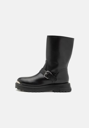 Stivale in pelle nera con punta arrotondata, cinturino regolabile con fibbia metallica e suola in gomma spessa e texturizzata. Dettaglio della punta in metallo argentato.