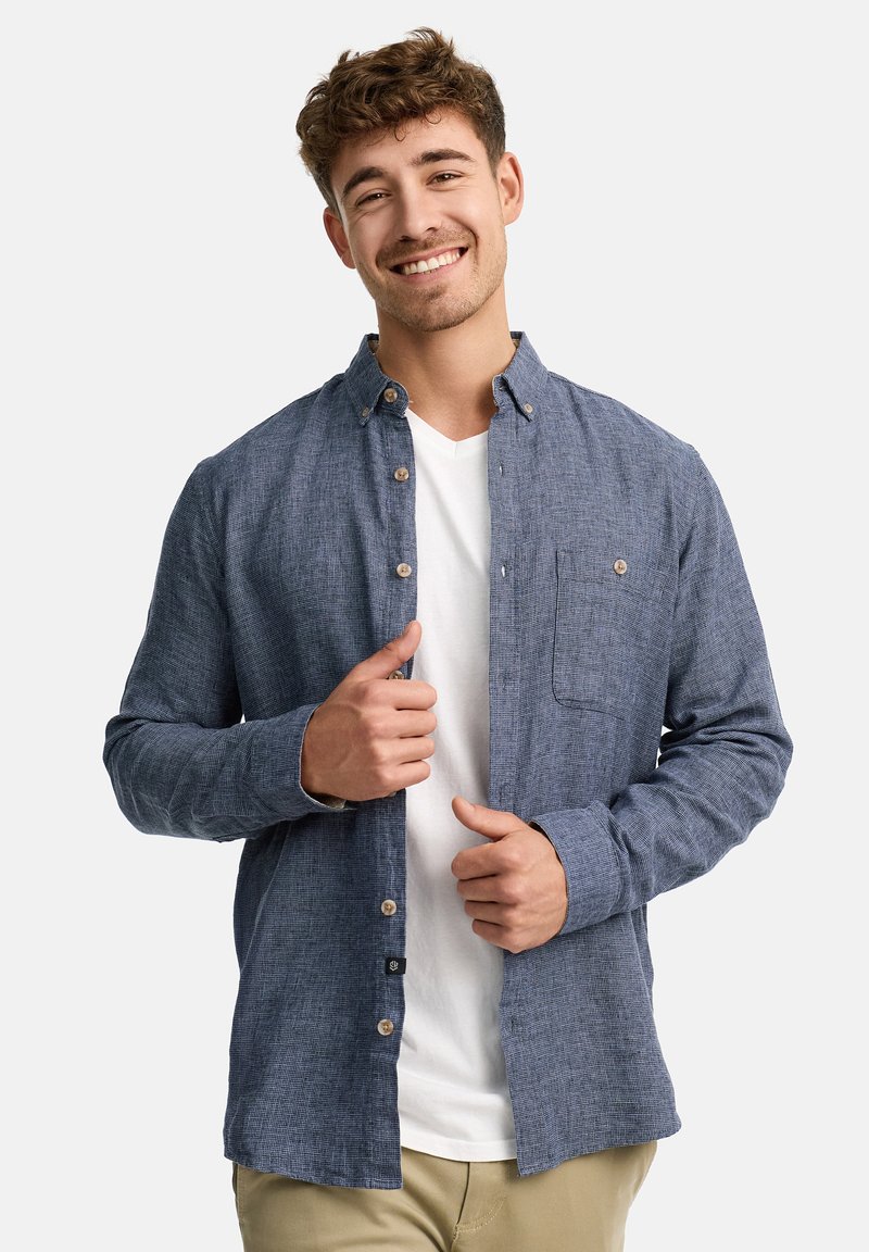 Camisa azul de botones con tejido texturizado, un bolsillo en el pecho y puños abotonados, superpuesta sobre una camiseta blanca. Corte delgado y ajustado.