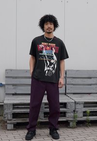Zwart oversized t-shirt met grafische print, ontspannen paarse broek en zwarte sneakers, staand op een houten bank tegen een grijze muur.