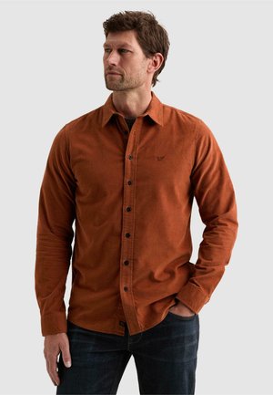 Homme aux cheveux courts et bruns portant une chemise boutonnée couleur rouille et un jean foncé, posant avec une main dans la poche sur un fond uni.