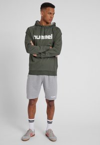 Olivgrön huvtröja med vitt logotyp, grå träningsshorts och grå sneakers med röda detaljer. Har vita ränder på ankelsockor.