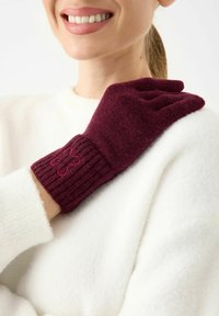 Gant de laine bordeaux avec poignet côtelé et détail brodé de fleur. La main du modèle repose sur un fond de pull blanc et duveteux.