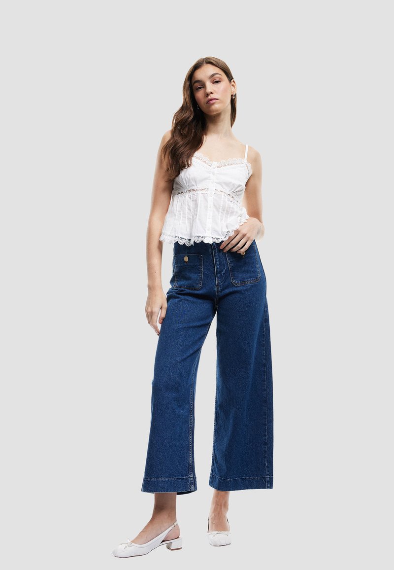 Vit blus med spetsdetaljer och knappar, matchad med högmidjade, vida mörkblå jeans och vita slingback-skor.