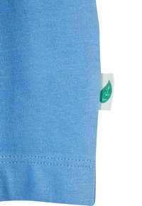 Tissu en coton bleu clair avec une texture lisse. Présente une petite étiquette blanche avec un motif de feuille verte près de l'ourlet. La couture est visible autour du bord.